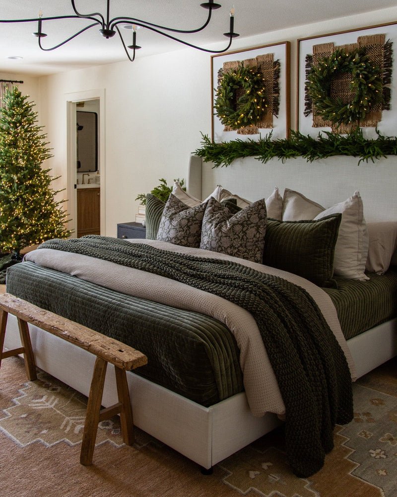 Bedroom with two lit wreaths over woven art, garland along the headboard, green velvet bedding, a knit throw and a Christmas tree