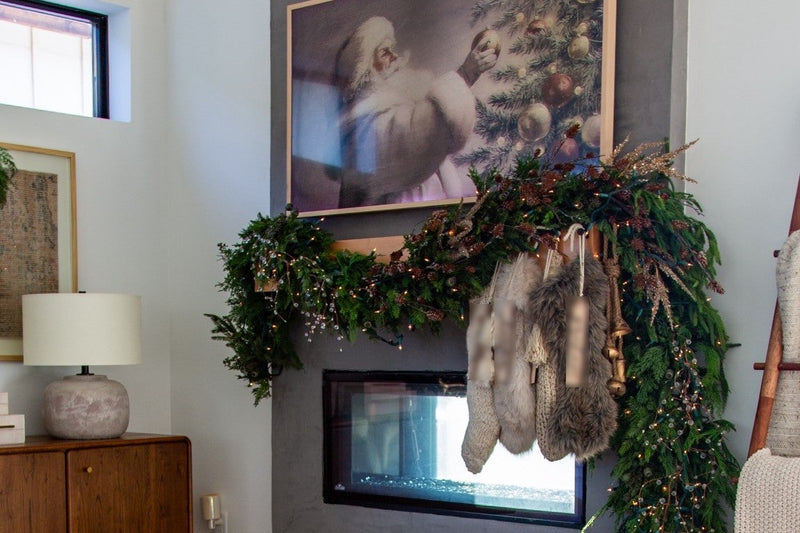 Christmas mantel layered with Norfolk pine and cedar garlands, pinecone stems and warm twinkle lights, with ivory and brown faux fur stockings and gold bells hanging below