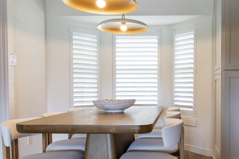 Dining nook with two brass pendant lights over a wood table, cream upholstered chairs and a white bowl in front of shuttered windows