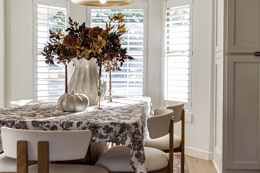Dining nook with a toile tablecloth, cream chairs, a fall floral centerpiece with white pumpkins and a brass pendant light