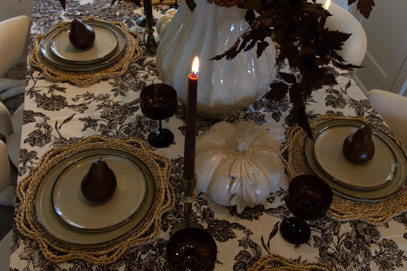 Close-up of a fall dinner table with woven chargers, stoneware plates, brown coupe glasses, a lit chocolate taper candle and a white ceramic pumpkin on a black-and-white toile tablecloth
