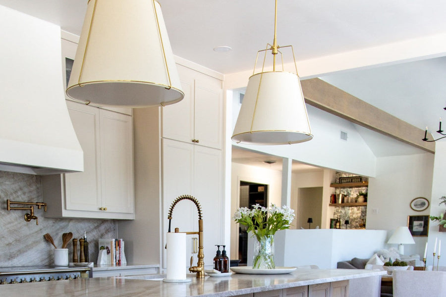 Builder-grade lighting swap in a kitchen: two oversized linen and brass pendant lights over a light wood island with stone counters