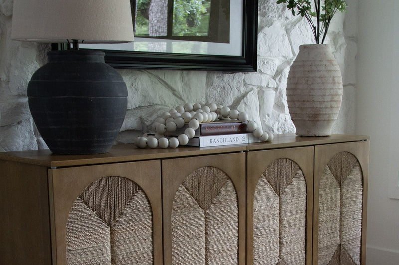 Woven-front wood sideboard in a living room with a black ceramic table lamp, a wood bead garland on stacked books and a cream vase of green branches