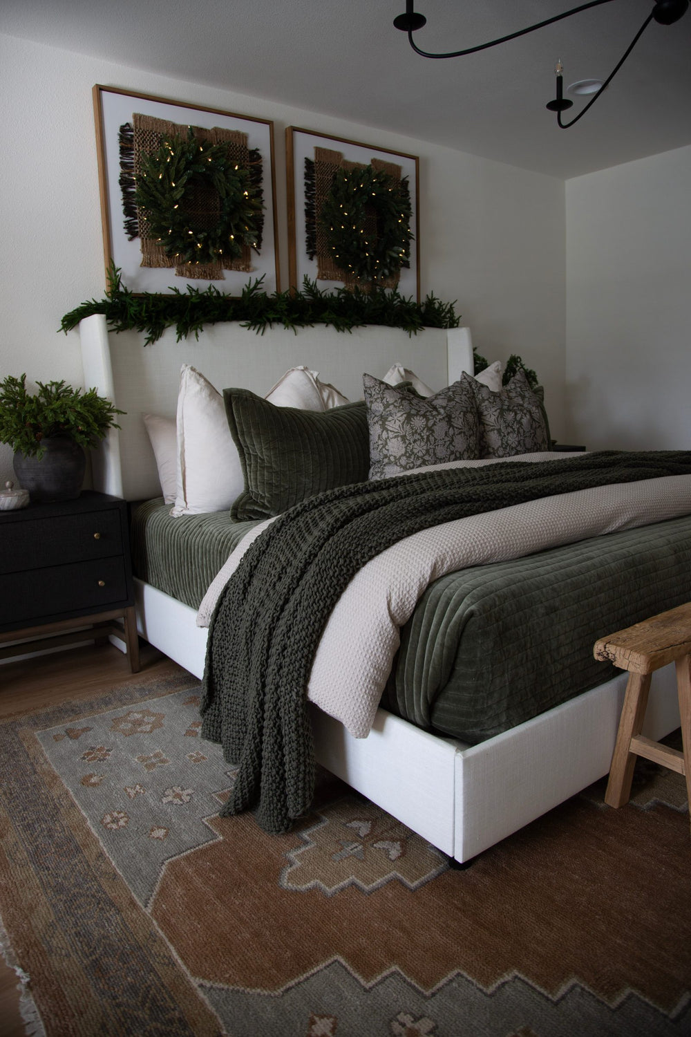 Bedroom with a white upholstered bed, green quilted coverlet and knit throw, framed wreaths with string lights and a patterned rug
