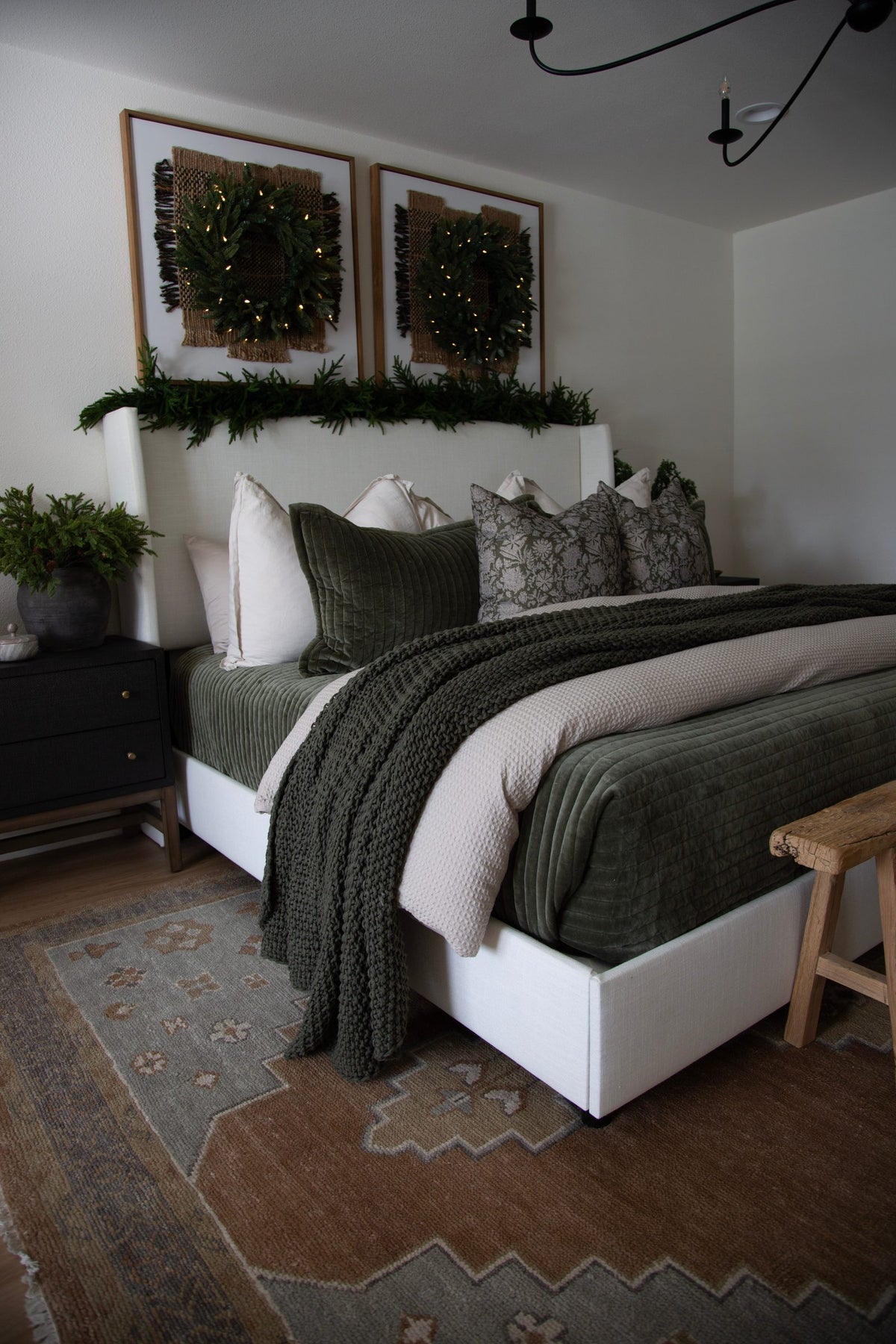 Bedroom with a white upholstered bed, green quilted coverlet and knit throw, framed wreaths with string lights and a patterned rug
