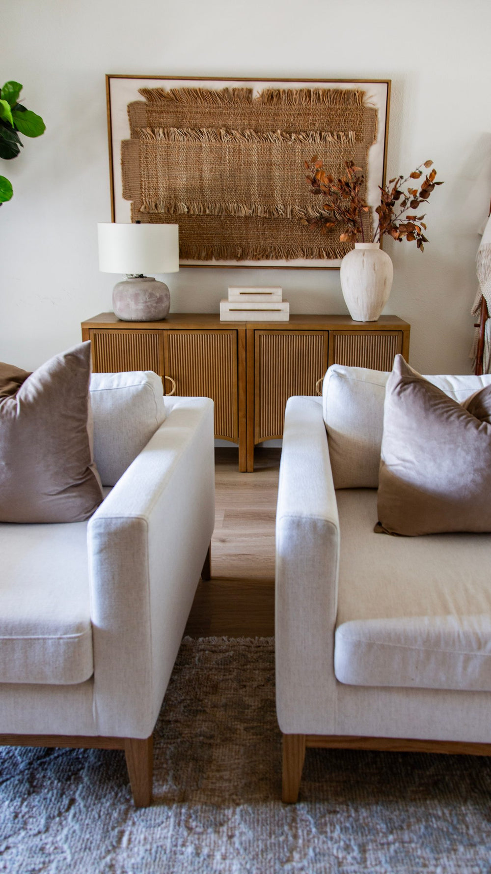 Two cream armchairs with taupe pillows facing a wood console with a lamp, vase and woven wall art