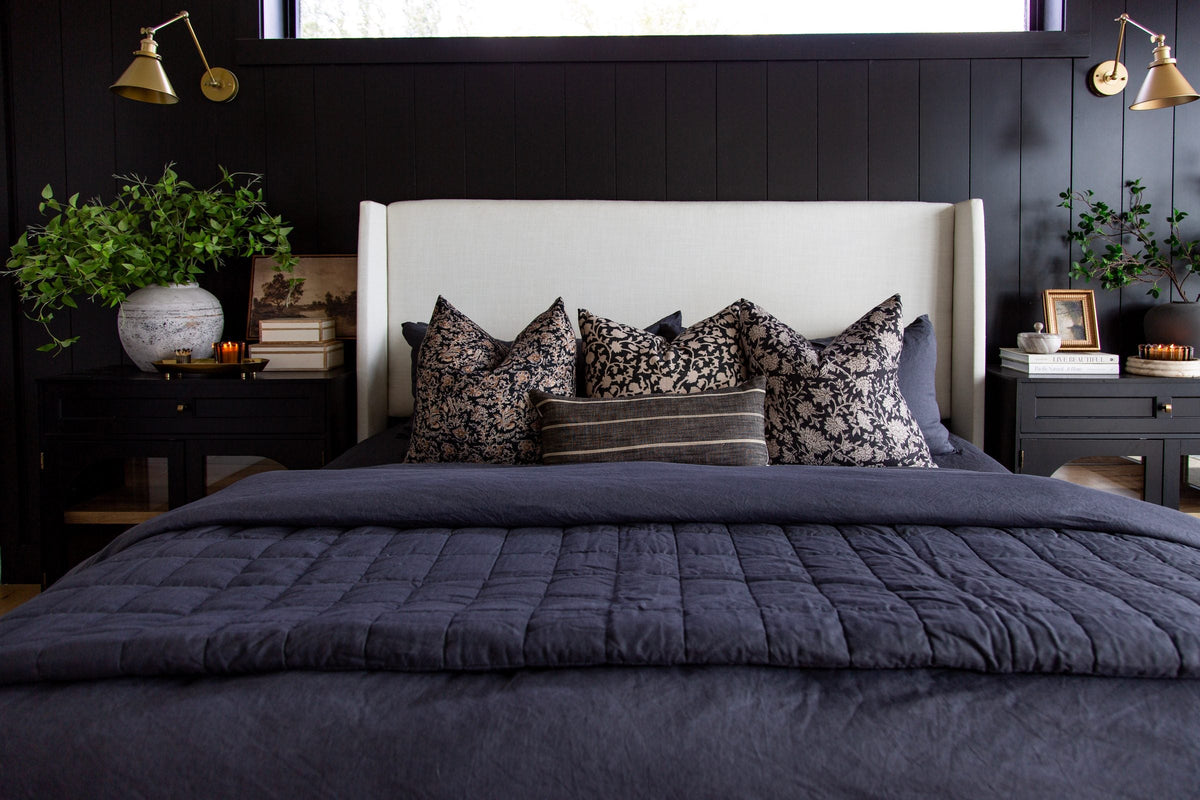 Bed with a navy quilt and floral pillows against a black vertical-panel wall with brass swing-arm sconces