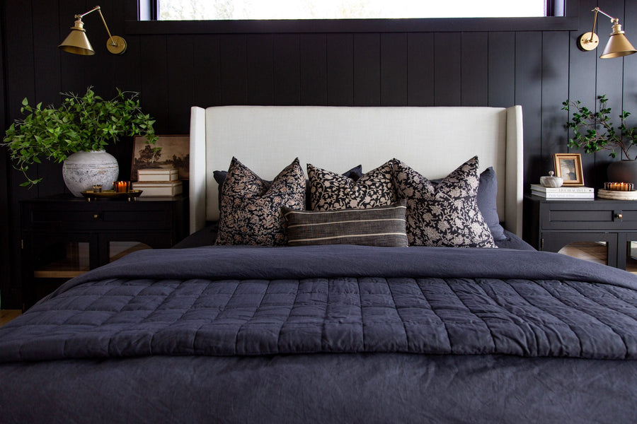 Bed with a navy quilt and floral pillows against a black vertical-panel wall with brass swing-arm sconces