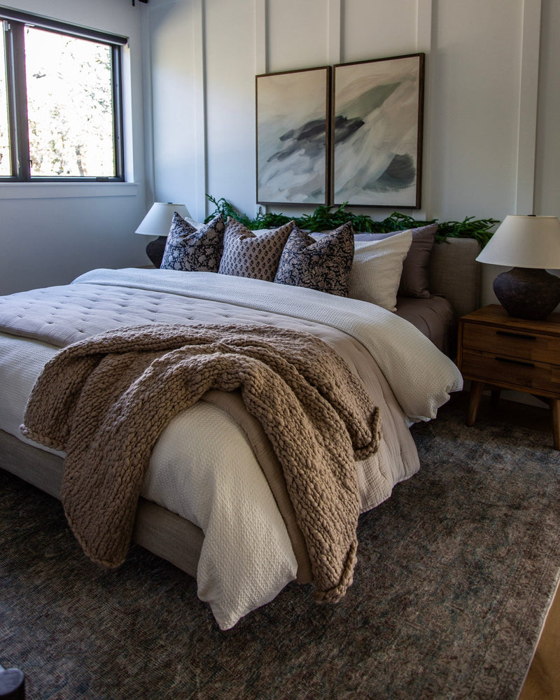 Bedroom with a white board-and-batten wall, layered cream and taupe bedding, a chunky knit throw and two abstract paintings