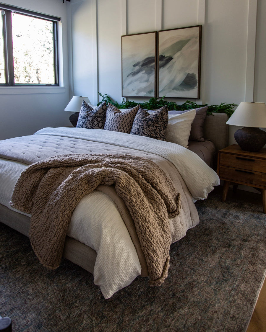 Bedroom with a white board-and-batten wall, layered cream and taupe bedding, a chunky knit throw and two abstract paintings