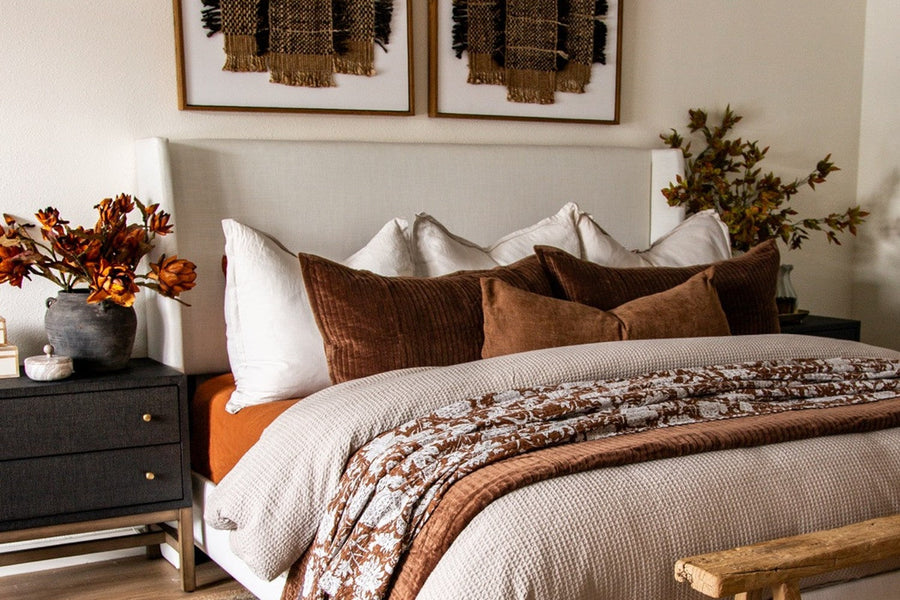 Ivory wingback upholstered bed with rust and brown velvet pillows, a floral quilt, a dark raffia and wood nightstand and fall stems