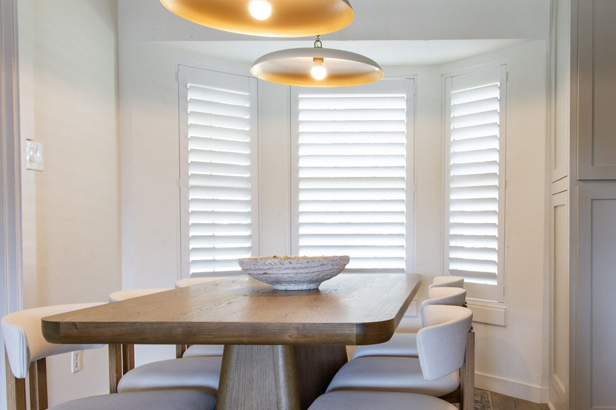 Dining nook with two brass pendant lights over a wood table, cream upholstered chairs and a white bowl in front of shuttered windows