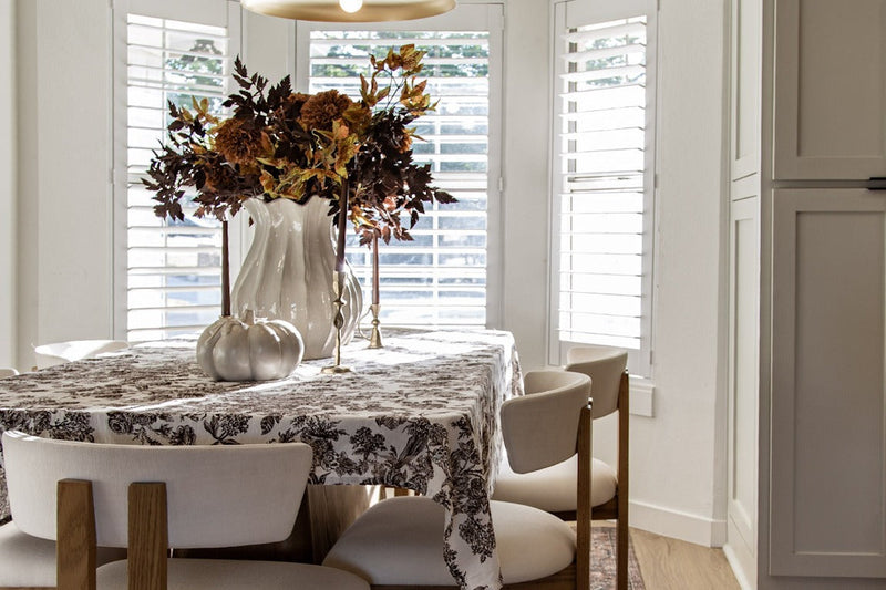 Dining nook with a toile tablecloth, cream chairs, a fall floral centerpiece with white pumpkins and a brass pendant light