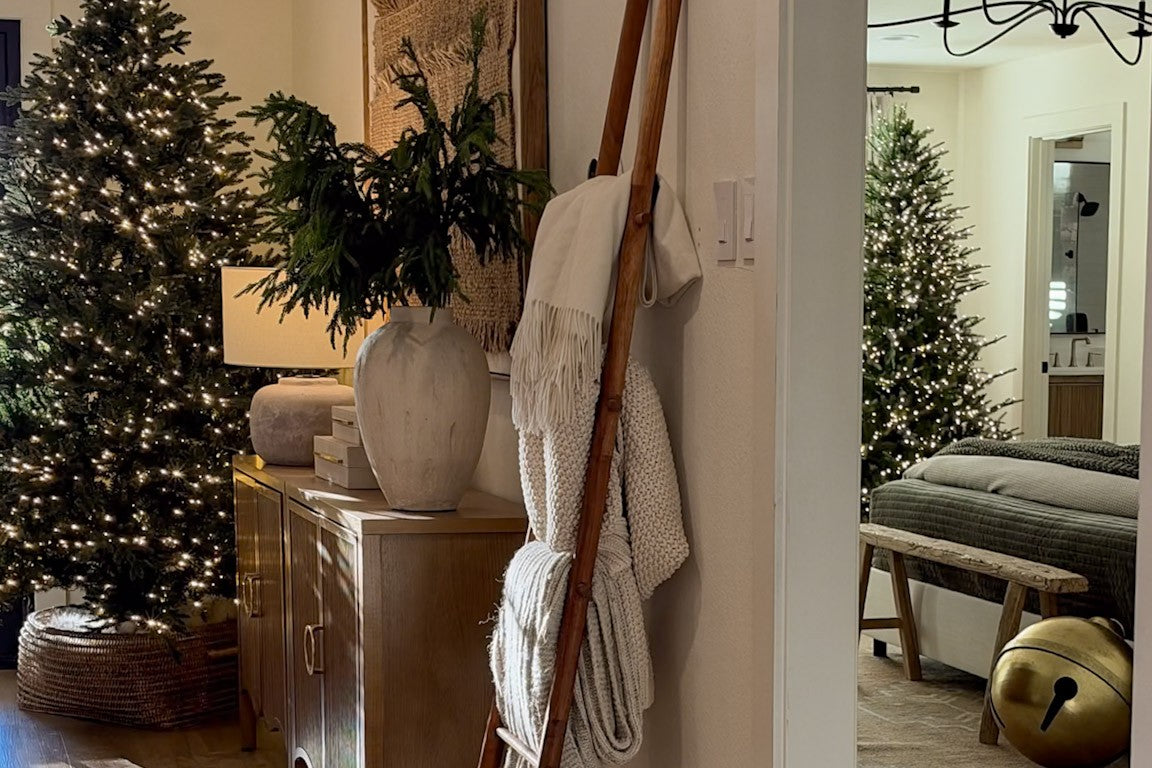 Entry hall dressed for the holidays with a lit Christmas tree in a woven basket, a wood console with greenery in a stone vase, and a wood blanket ladder