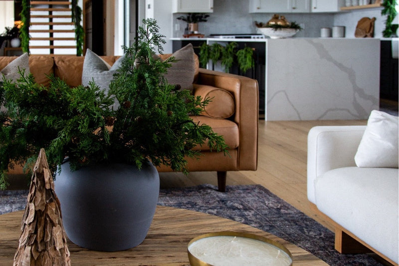 Budget Christmas decor in a living room: cedar and pinecone greenery in a slate vase, a carved wood tree and a brass candle on a round wood coffee table
