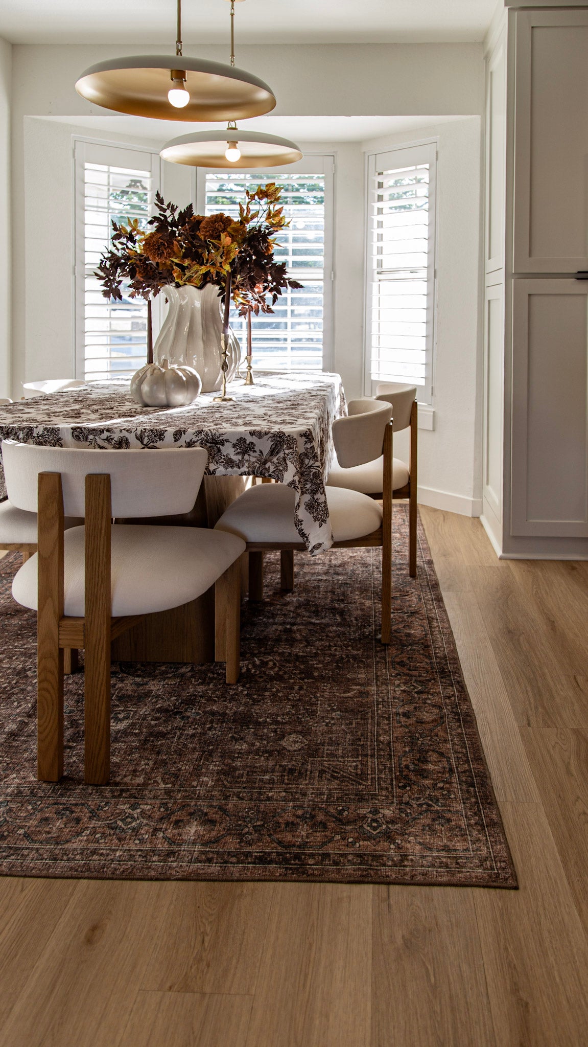 Dining nook with toile tablecloth, cream chairs, fall floral centerpiece and two brass pendant lights