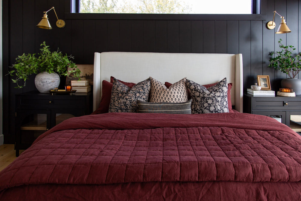 Bed with a burgundy quilt and patterned pillows against a black vertical-panel wall with brass swing-arm sconces