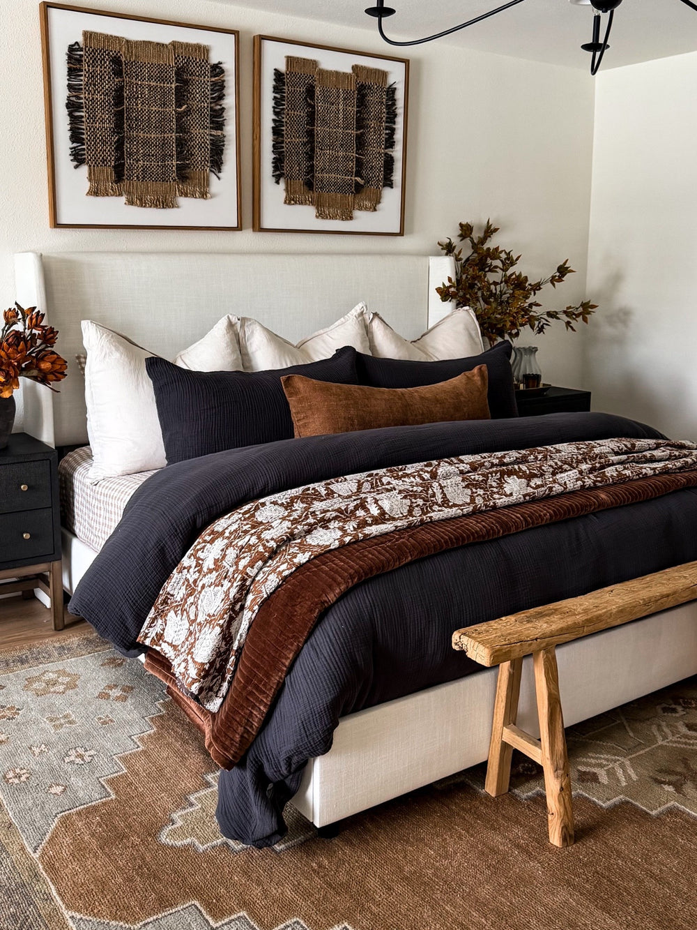 Bedroom with a white upholstered bed in charcoal and chestnut bedding, a floral quilt, navy and rust pillows, a wood bench, woven wall art and a patterned rug