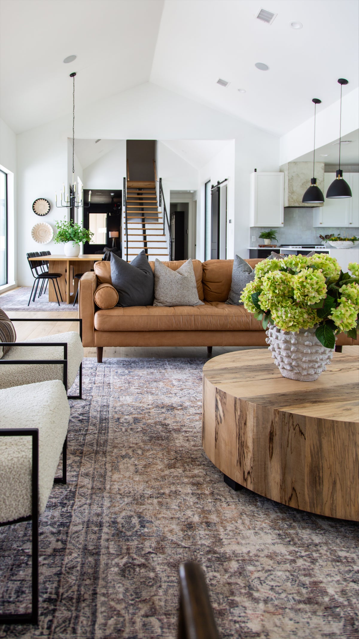 Open living room with a cognac leather sofa, gray and oatmeal pillows, green hydrangeas in a textured white vase and a round wood coffee table on a vintage-style rug, with a staircase and dining table beyond
